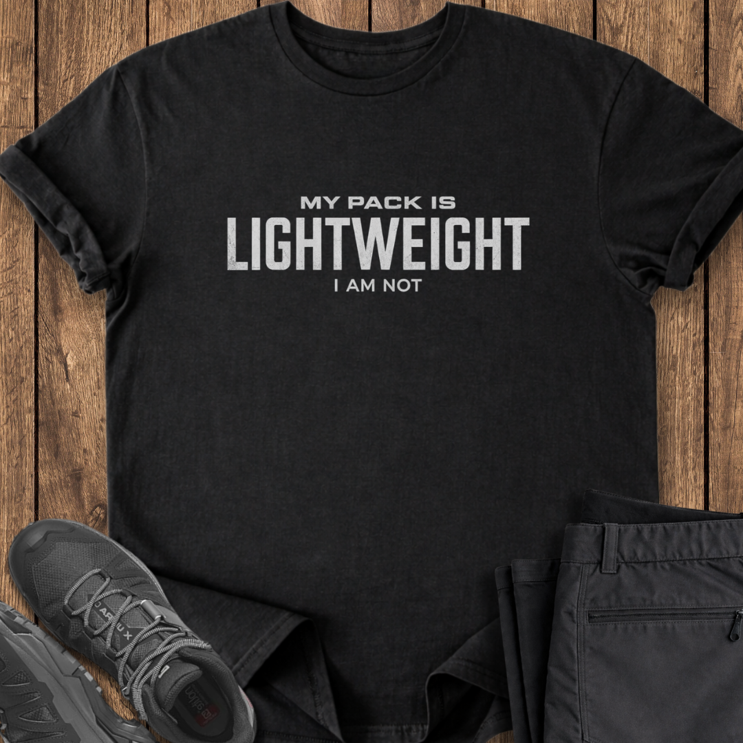 Black t-shirt with 'My pack is lightweight, I am not' text on a wooden surface with shoes and pants. backpacking funny t shirt