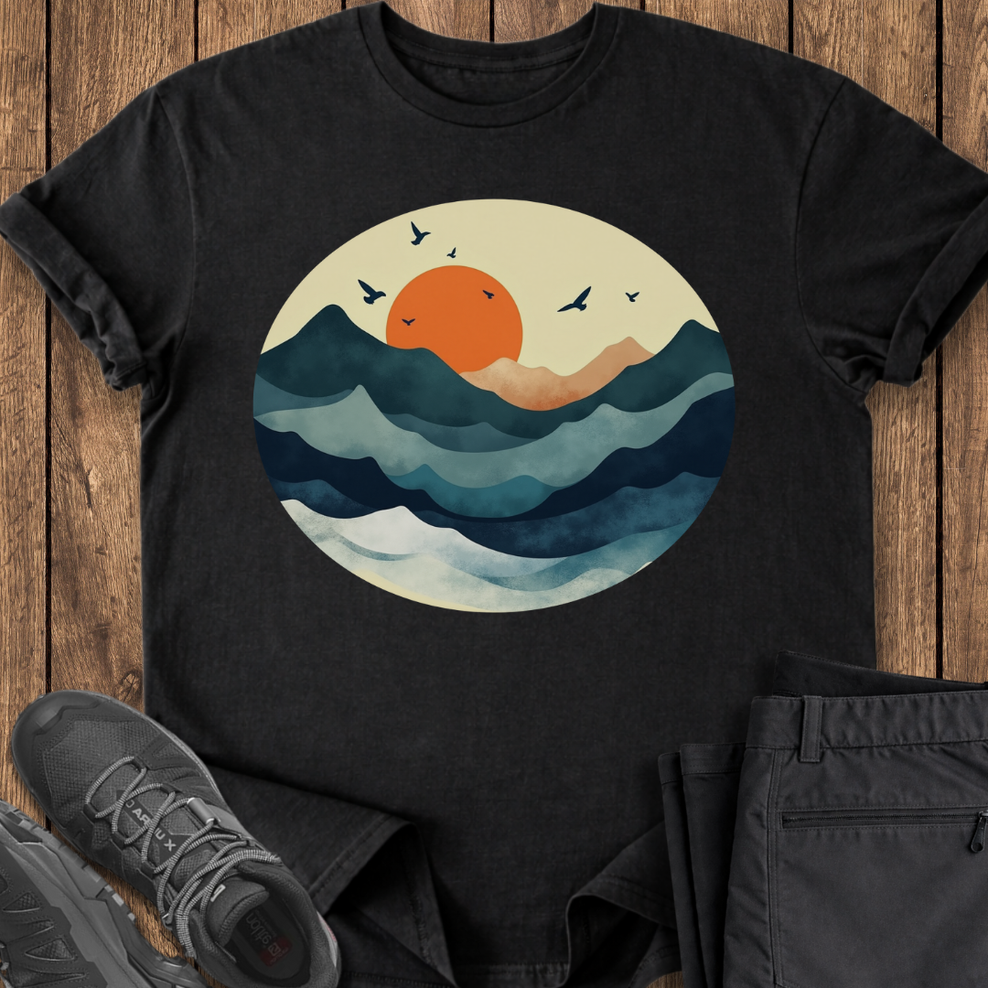 Black t-shirt with a sunset and wave design on a wooden surface with shoes and pants.