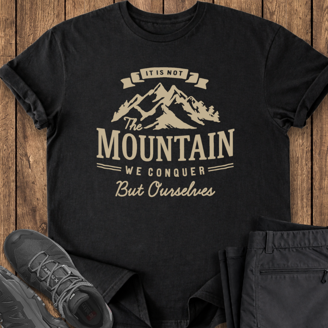 Black t-shirt with mountain graphic and text on a wooden surface with shoes and pants.