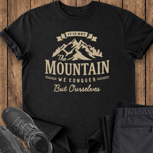 Black t-shirt with mountain graphic and text on a wooden surface with shoes and pants.