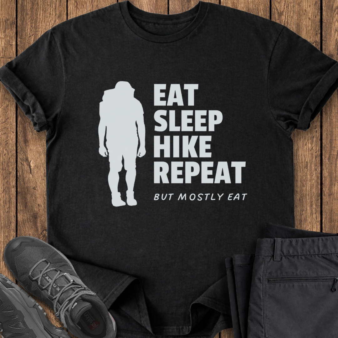 Black t-shirt with 'Eat Sleep Hike Repeat' text and hiking silhouette on a wooden surface with shoes and pants. funny hiking shirt