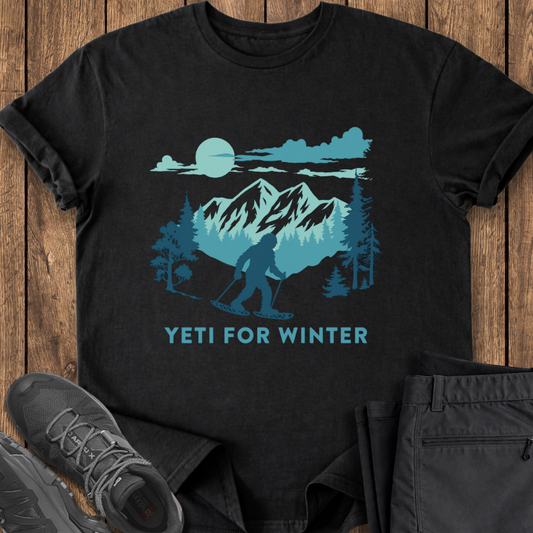 Black t-shirt with blue graphic of a yeti and mountains, text 'YETI FOR WINTER', on a wooden surface with shoes and pants.