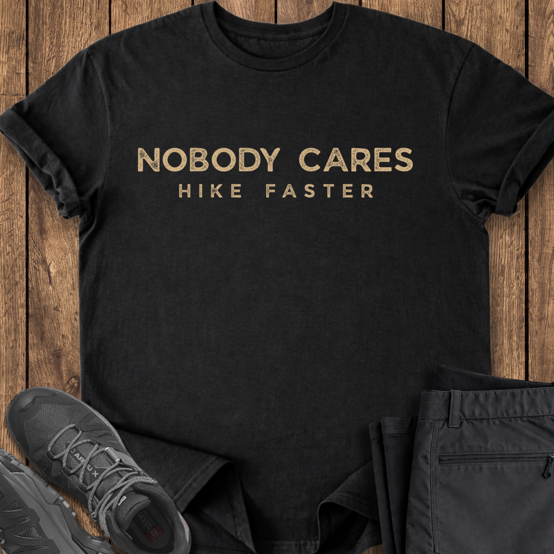 Black t-shirt with 'Nobody Cares Hike Faster' text on a wooden surface with shoes and pants.