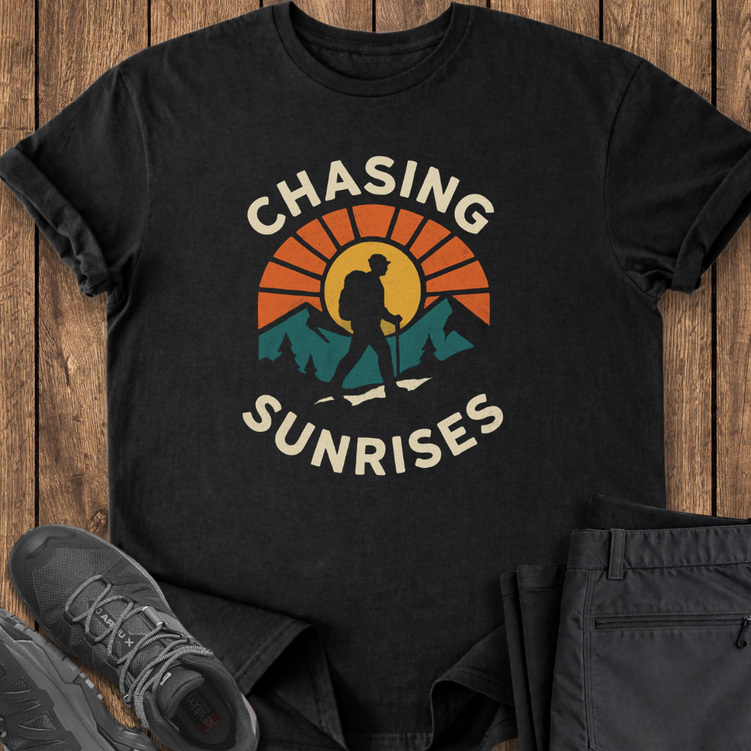 Black t-shirt with 'Chasing Sunrises' design on a wooden surface with shoes and pants.