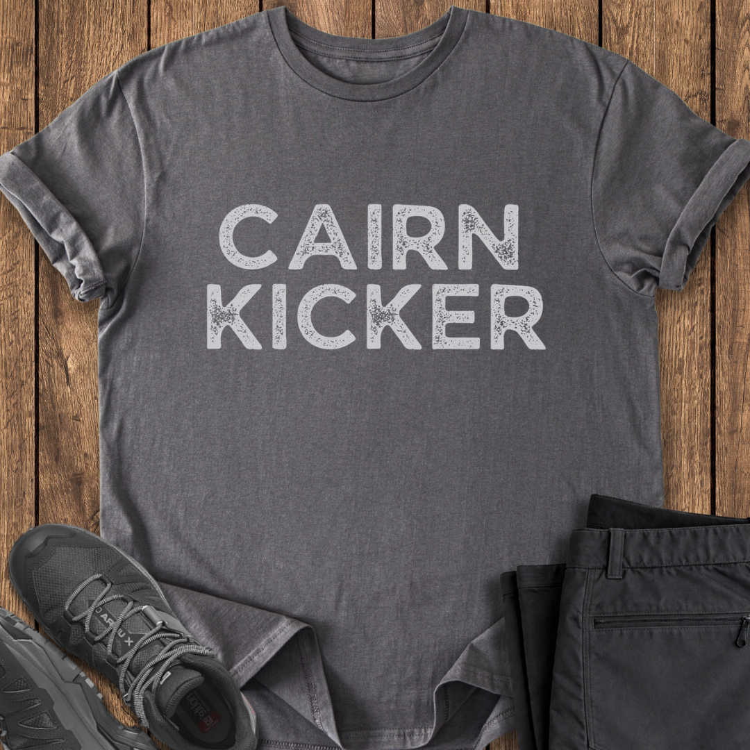 Gray t-shirt with 'CAIRN KICKER' text on a wooden surface with shoes and pants. hiking t shirt