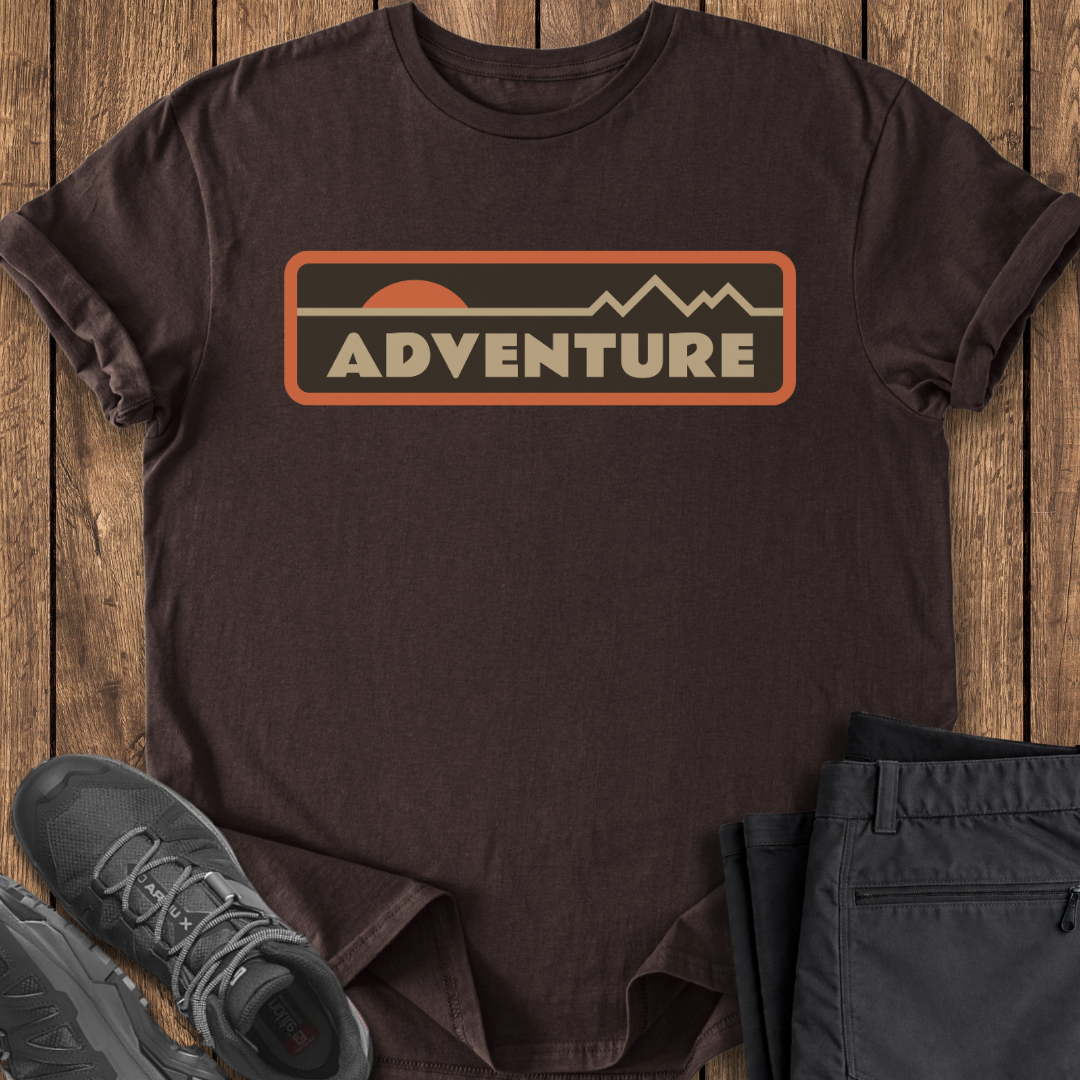 Brown t-shirt with 'ADVENTURE' graphic on a wooden surface with shoes and pants.