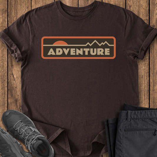 Brown t-shirt with 'ADVENTURE' graphic on a wooden surface with shoes and pants.