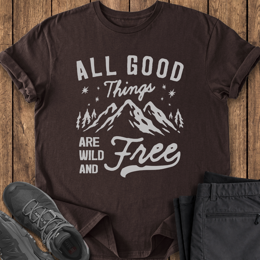 Brown t-shirt with 'All Good Things Are Wild and Free' text and mountain graphic on a wooden surface with shoes and pants.