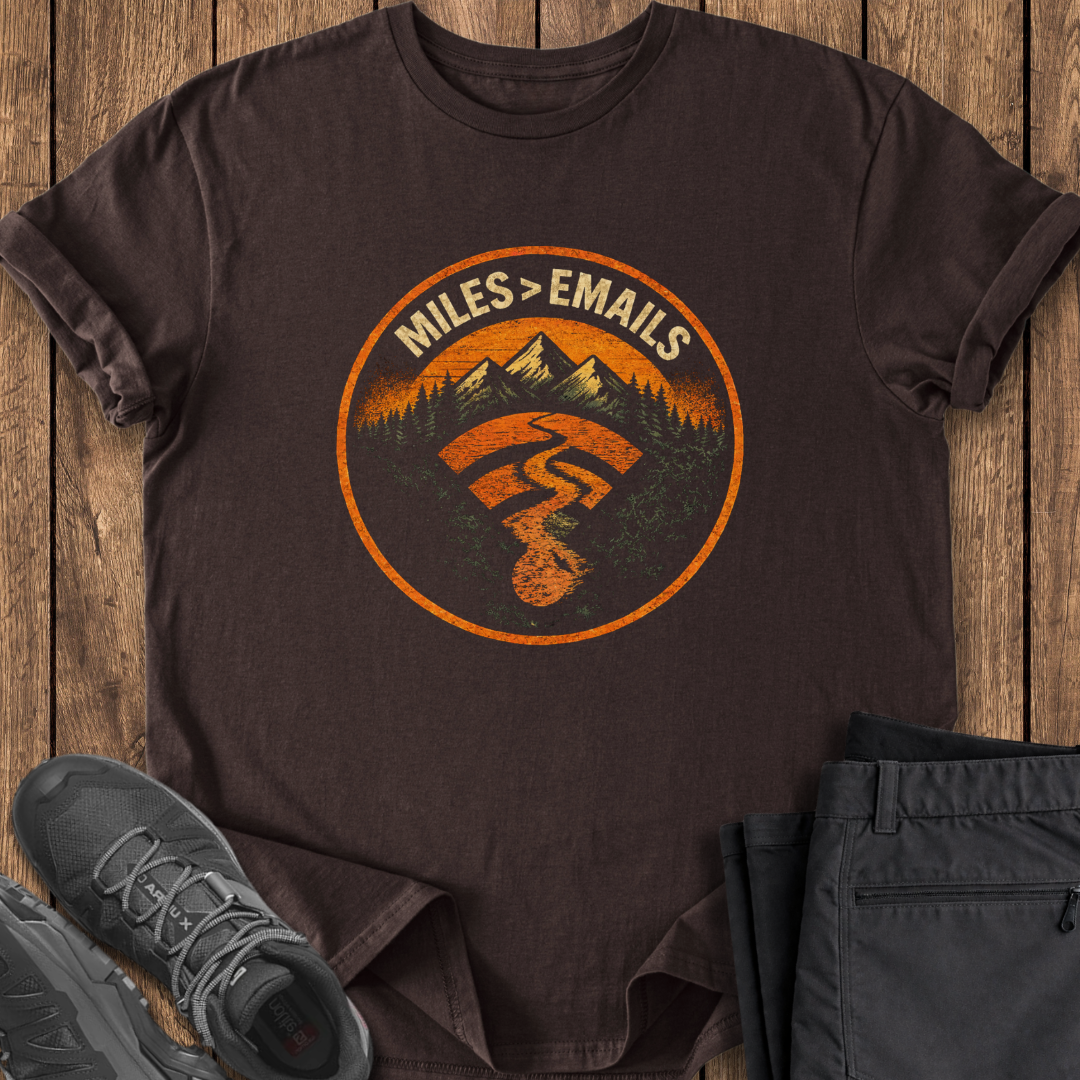 Brown t-shirt with a graphic design featuring mountains and text, placed on a wooden surface with shoes and pants. hiking t shirt