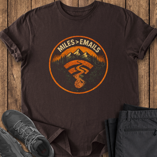 Brown t-shirt with a graphic design featuring mountains and text, placed on a wooden surface with shoes and pants. hiking t shirt