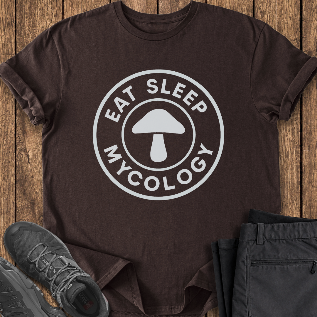 Brown t-shirt with 'Eat Sleep Mycology' design on a wooden surface with shoes and pants. mushroom foraging t shirt