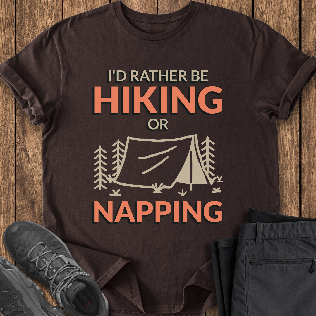Brown t-shirt with 'I'd Rather Be Hiking or Napping' text and tent graphic on a wooden surface with shoes and pants. funny camping hiking t shirt