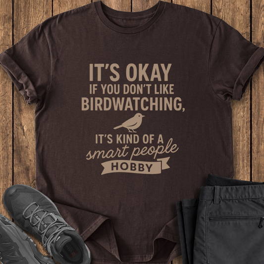 Brown t-shirt with humorous text and bird graphic on a wooden surface with shoes and pants.