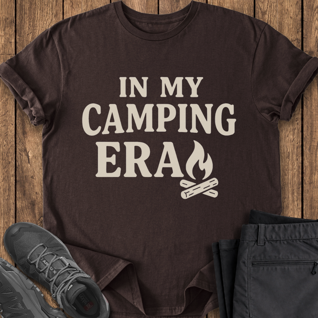 Brown t-shirt with 'In My Camping Era' text and campfire graphic on a wooden surface with shoes and pants. camp camper t shirt