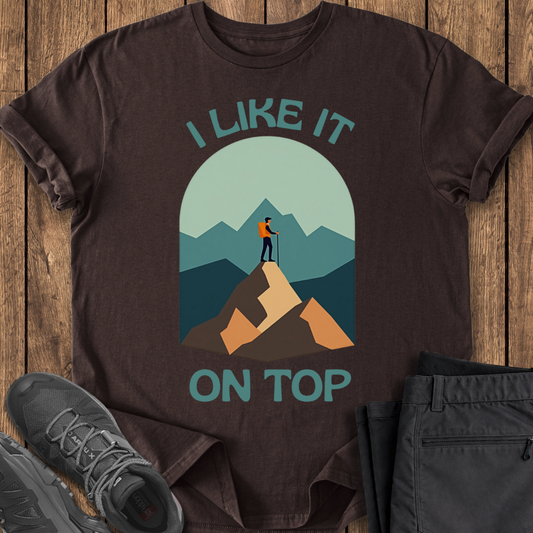 Brown t-shirt with mountain graphic and text 'I Like It On Top' on a wooden surface with shoes and pants.