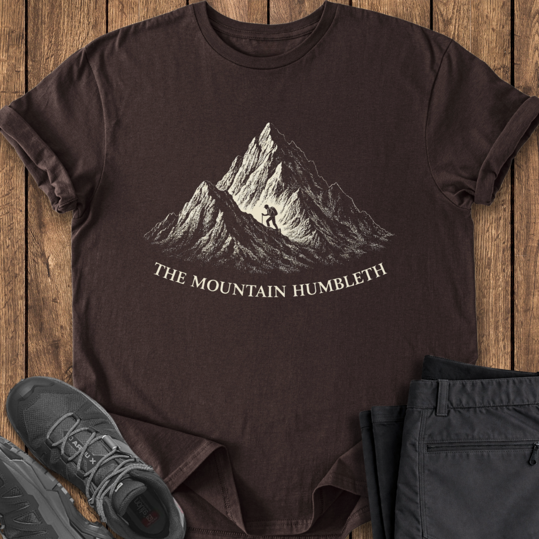 Brown t-shirt with mountain graphic and text on a wooden surface, accompanied by black pants and shoes. funny mountain hiking t shirt