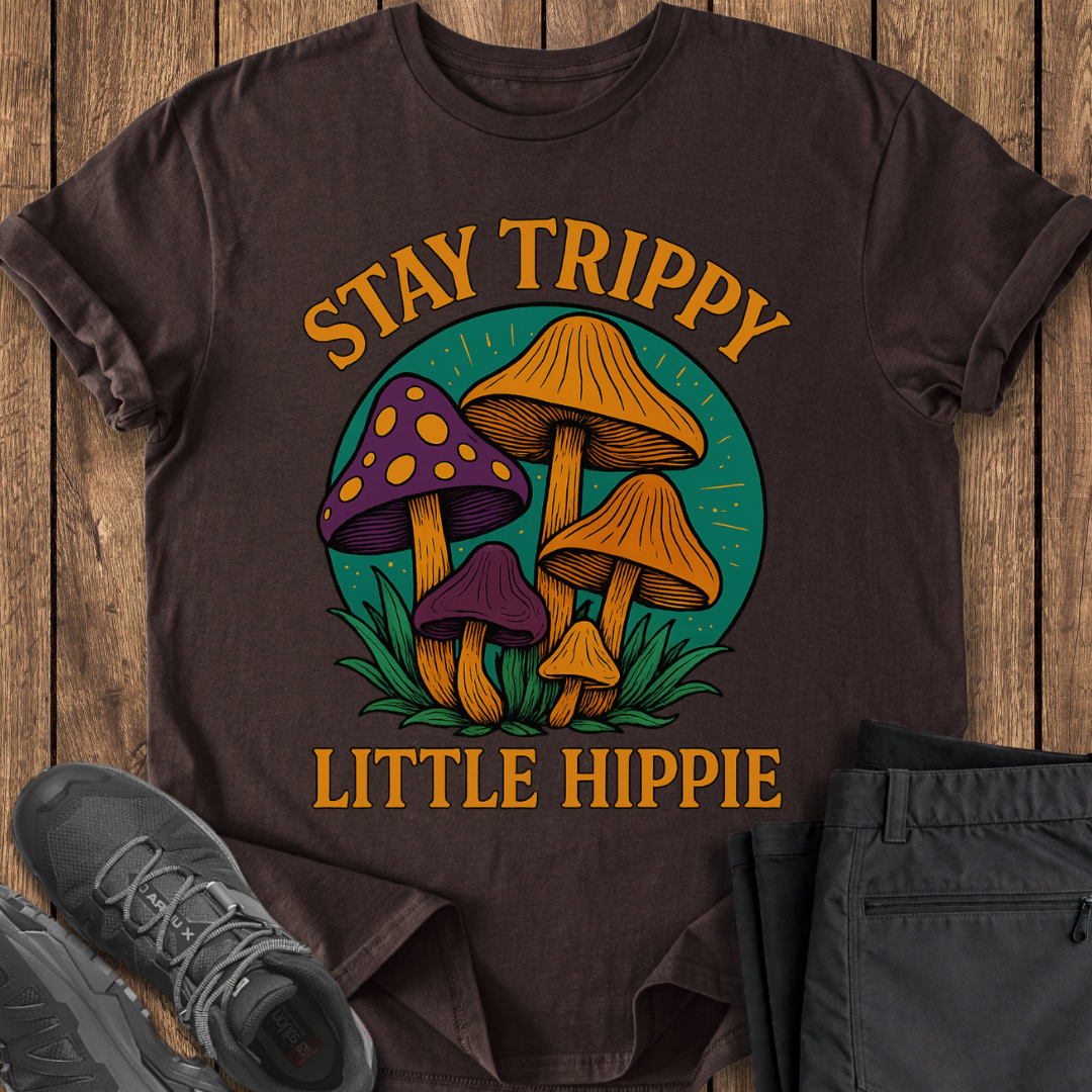 Brown t-shirt with mushroom graphic and text 'Stay Trippy Little Hippie' on a wooden surface.