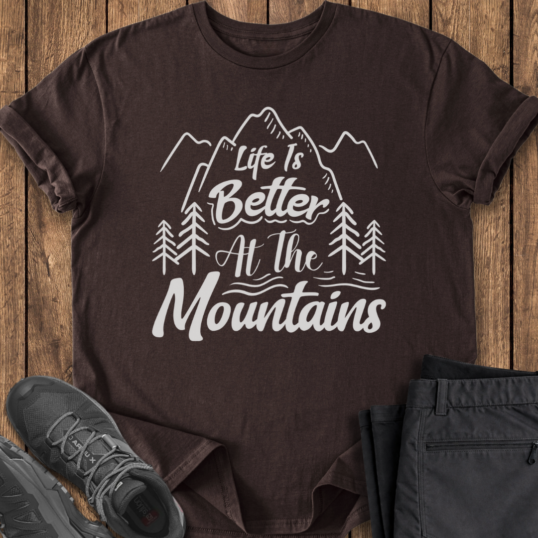 Brown t-shirt with 'Life is Better at the Mountains' design on a wooden surface with shoes and pants.