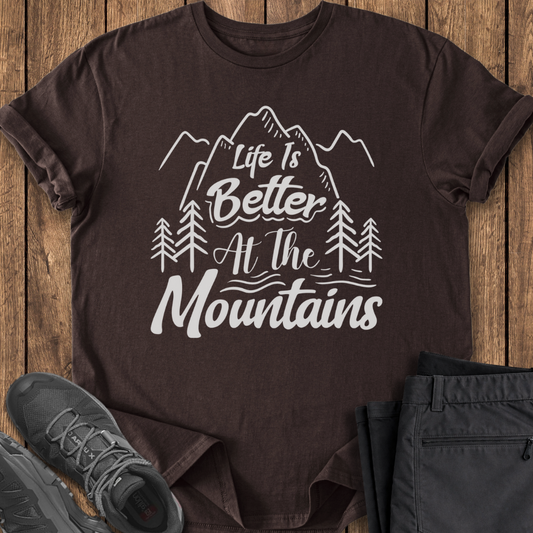 Brown t-shirt with 'Life is Better at the Mountains' design on a wooden surface with shoes and pants.