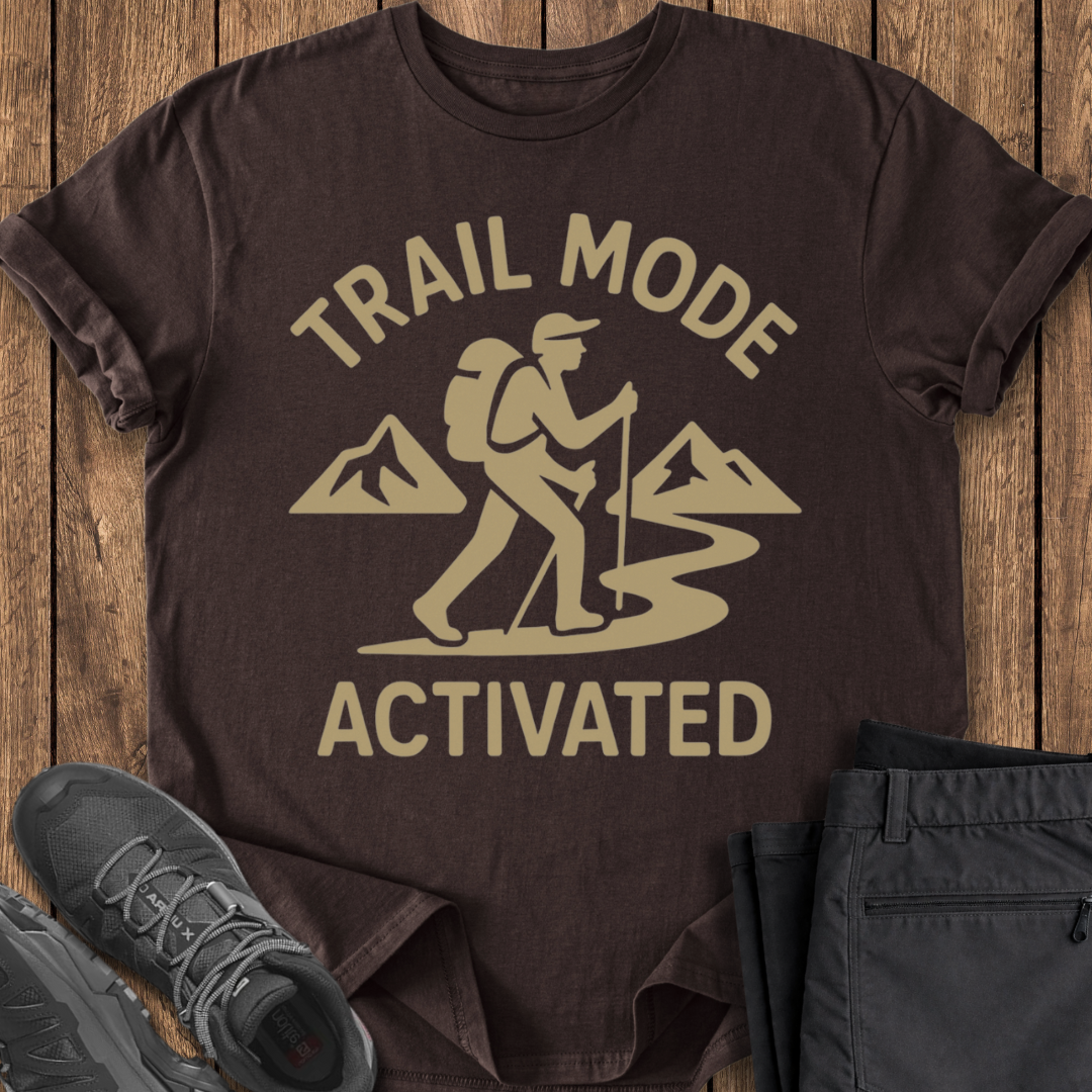 Brown t-shirt with 'Trail Mode Activated' graphic on a wooden surface with shoes and pants.