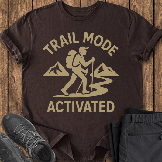 Brown t-shirt with 'Trail Mode Activated' graphic on a wooden surface with shoes and pants.