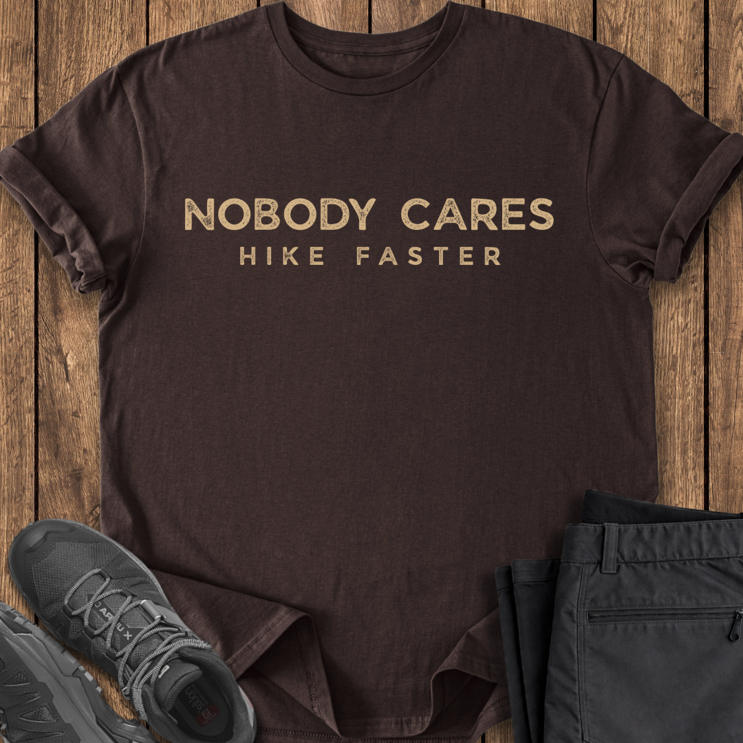 Brown t-shirt with 'Nobody Cares Hike Faster' text on a wooden surface with shoes and pants.