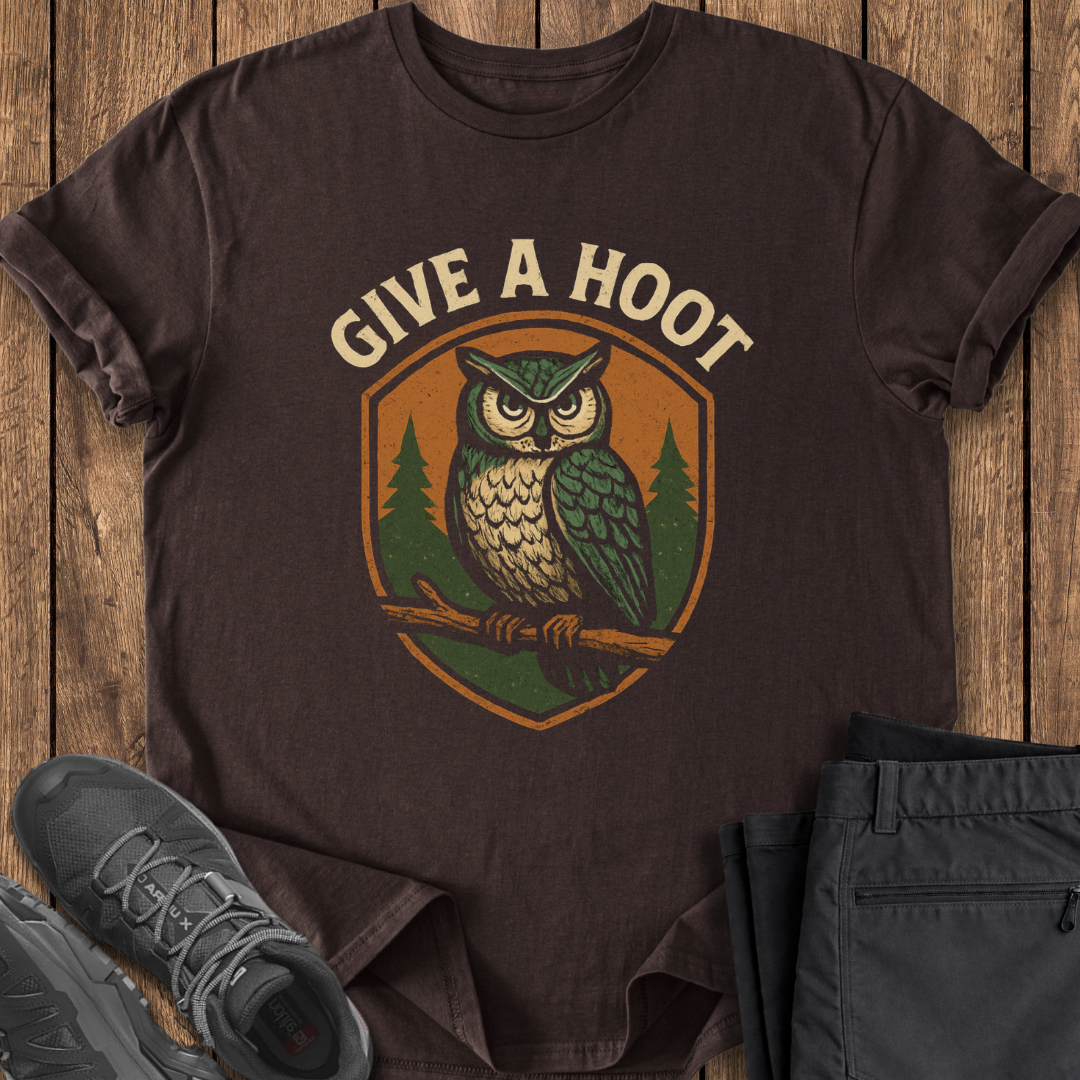 Brown t-shirt with owl graphic and 'Give a Hoot' text on a wooden surface with shoes and pants.