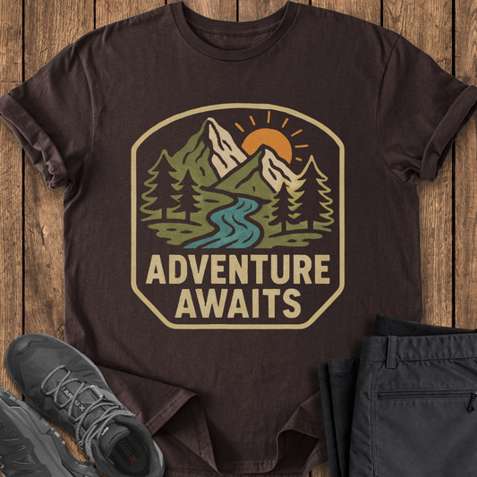 Brown t-shirt with 'Adventure Awaits' graphic on a wooden surface with shoes and pants. adventure mountain t shirt