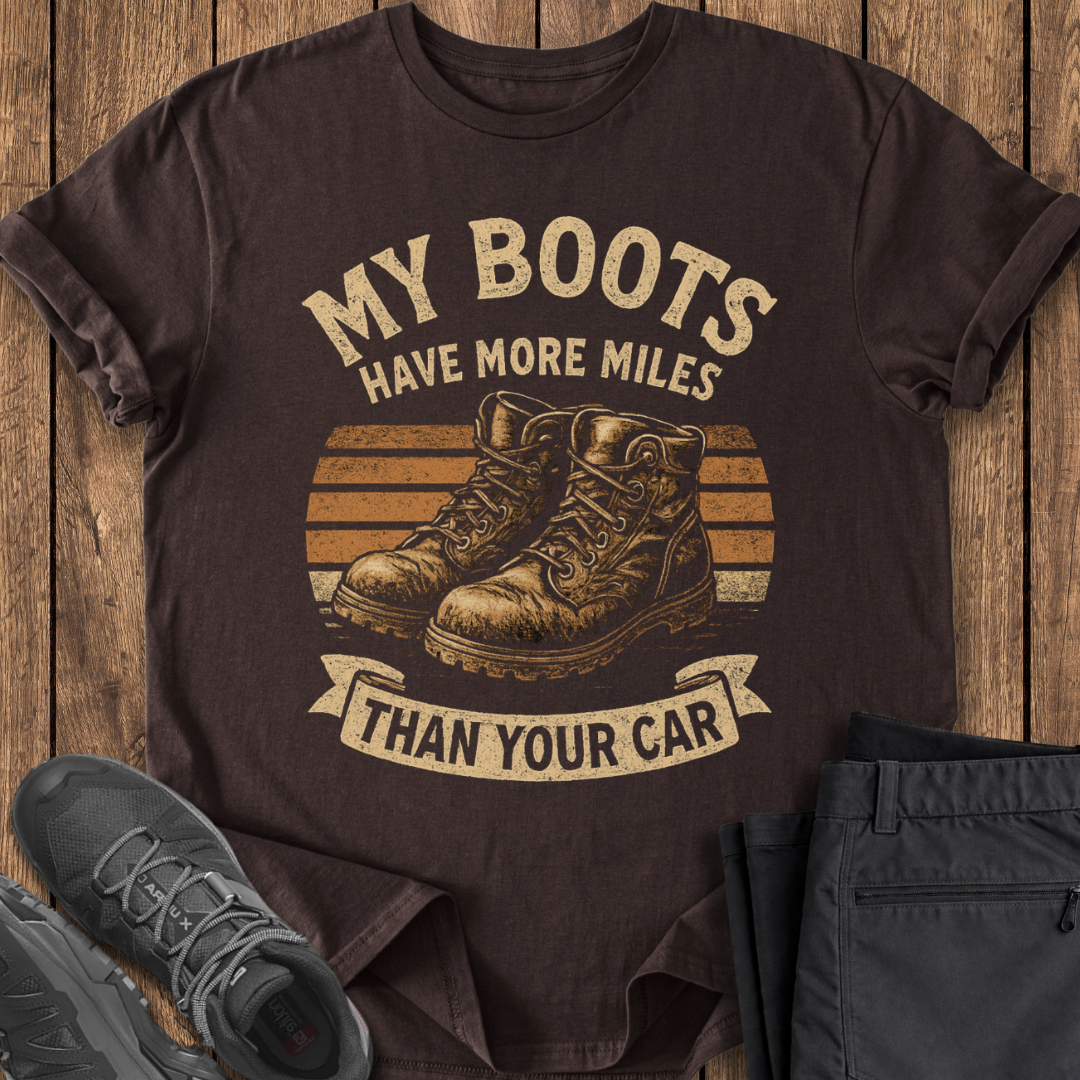 Brown t-shirt with graphic and text 'My boots have more miles than your car' on a wooden surface.