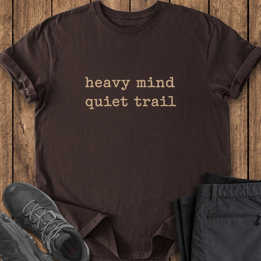 Brown t-shirt with 'heavy mind quiet trail' text on a wooden surface with shoes and pants. mental health hike shirt