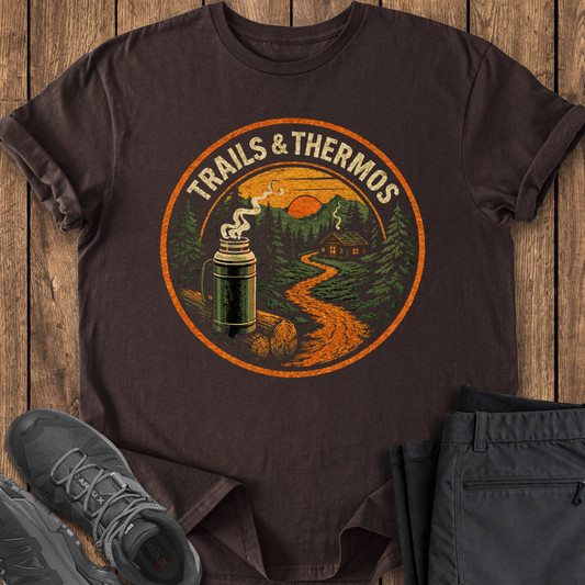Brown t-shirt with 'Trails & Thermos' graphic on a wooden surface with shoes and pants. hiking shirt