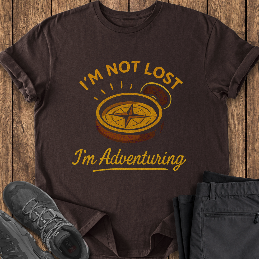 Brown t-shirt with 'I'm not lost, I'm adventuring' vintage compass design on a wooden surface with shoes and pants. adventure t shirt