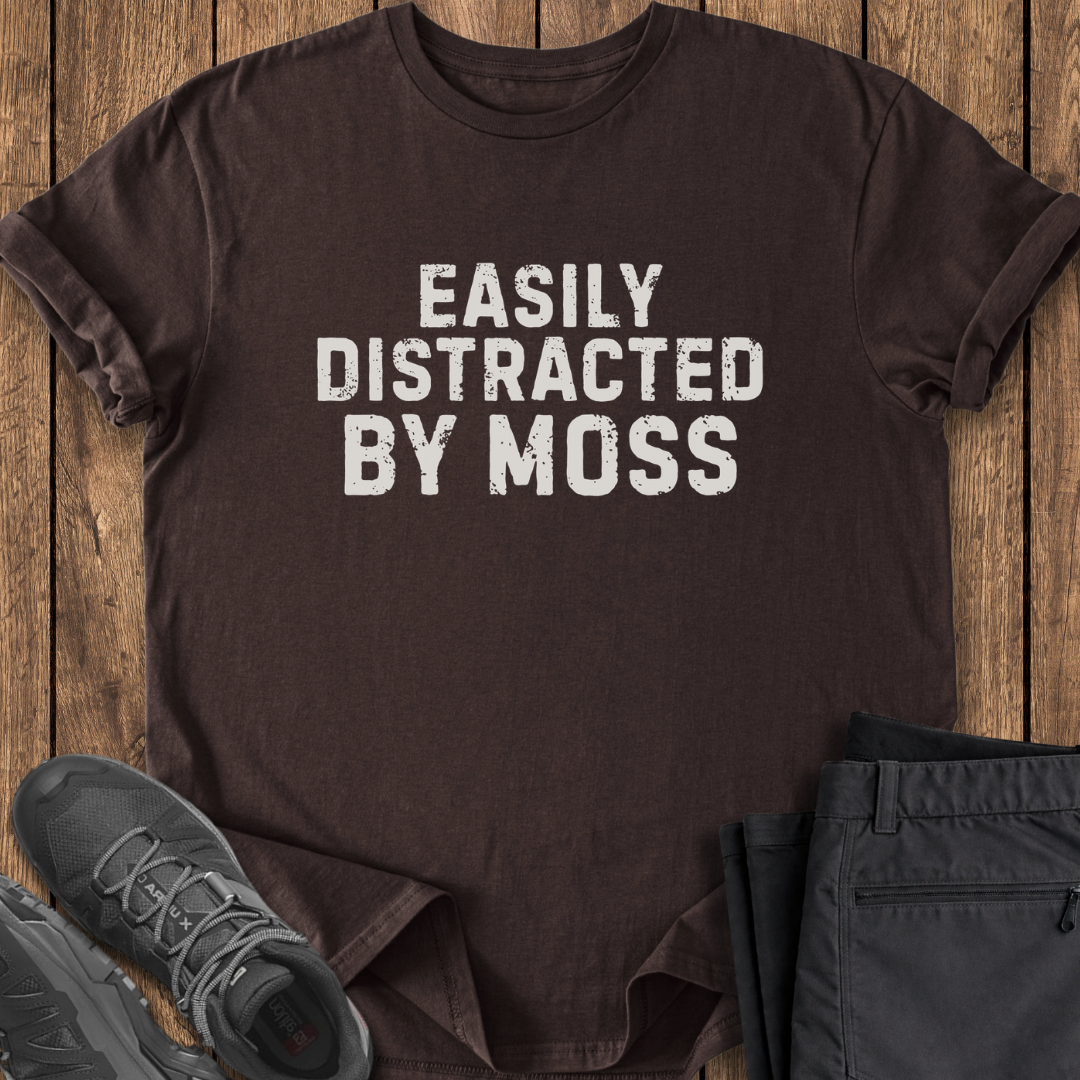 Brown t-shirt with 'Easily Distracted by Moss' text on a wooden surface with shoes and pants. funny hiking t shirt