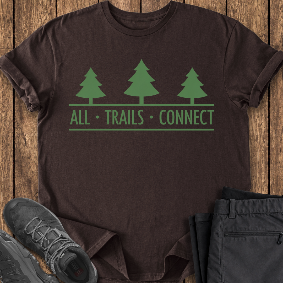 Brown t-shirt with green tree design and 'All Trails Connect' text on a wooden surface, with shoes and pants partially visible.
