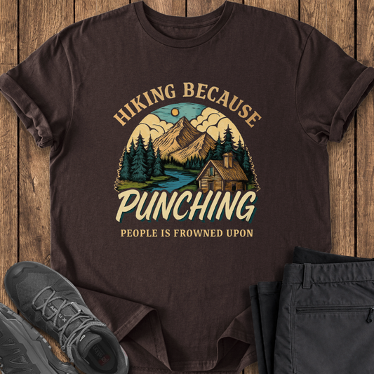 Brown t-shirt with hiking graphic and text on a wooden surface with shoes and pants. punching is frowned upon funny hiking shirt
