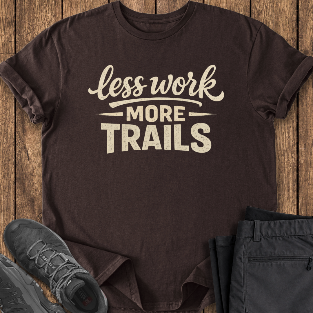 Brown t-shirt with 'less work more trails' text on a wooden surface with shoes and pants.