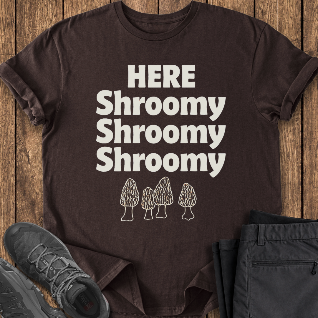 Brown t-shirt with 'HERE Shroomy Shroomy Shroomy' text and mushroom graphics on a wooden surface.