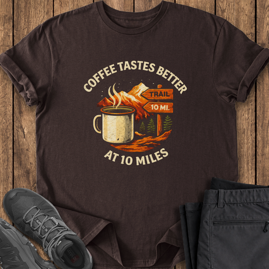 Brown t-shirt with coffee-themed graphic on a wooden surface with shoes and pants. coffee on the trail shirt