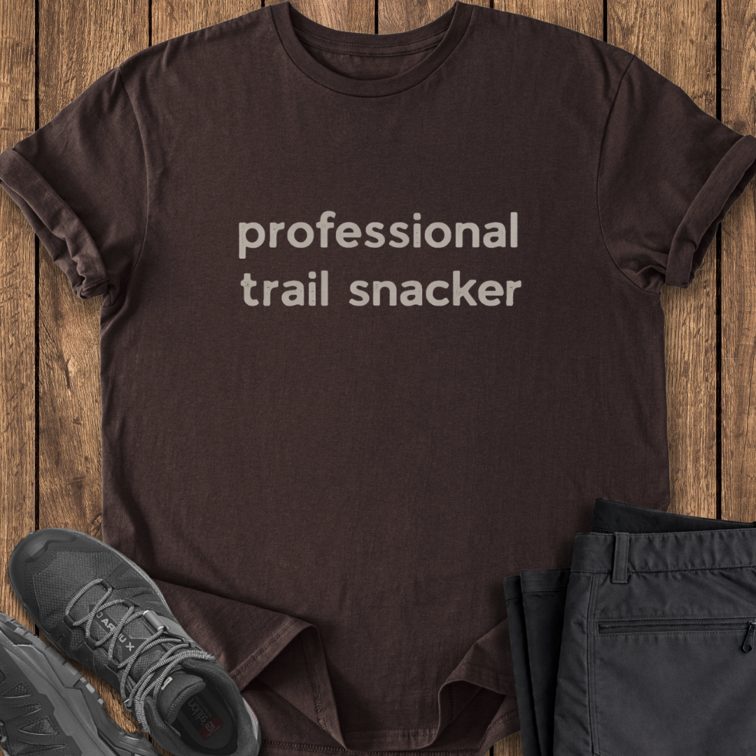 Brown t-shirt with 'professional trail snacker' text on a wooden surface with shoes and shorts. funny hike t shirt