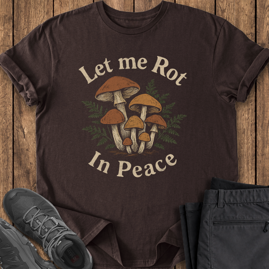 Brown t-shirt with mushroom design and text 'Let me Rot In Peace' on a wooden surface.