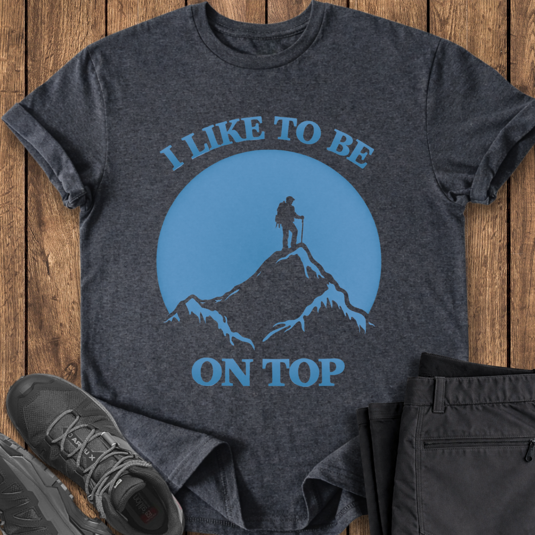 Dark gray t-shirt with blue graphic and text 'I LIKE TO BE ON TOP' on a wooden surface, with shoes and pants partially visible. funny hiking t shirt