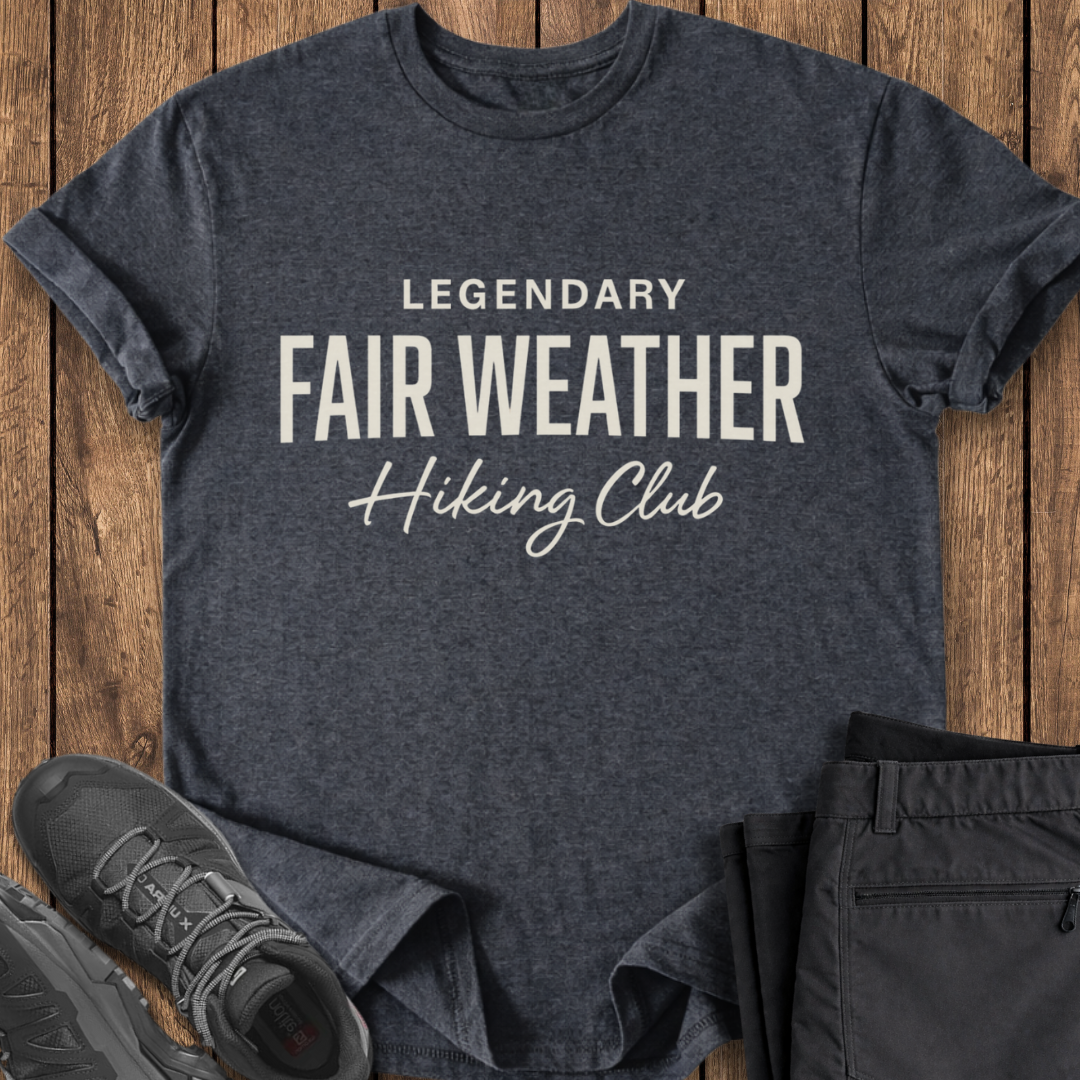 Dark gray t-shirt with 'Legendary Fair Weather Hiking Club' text on a wooden surface with black pants and shoes.