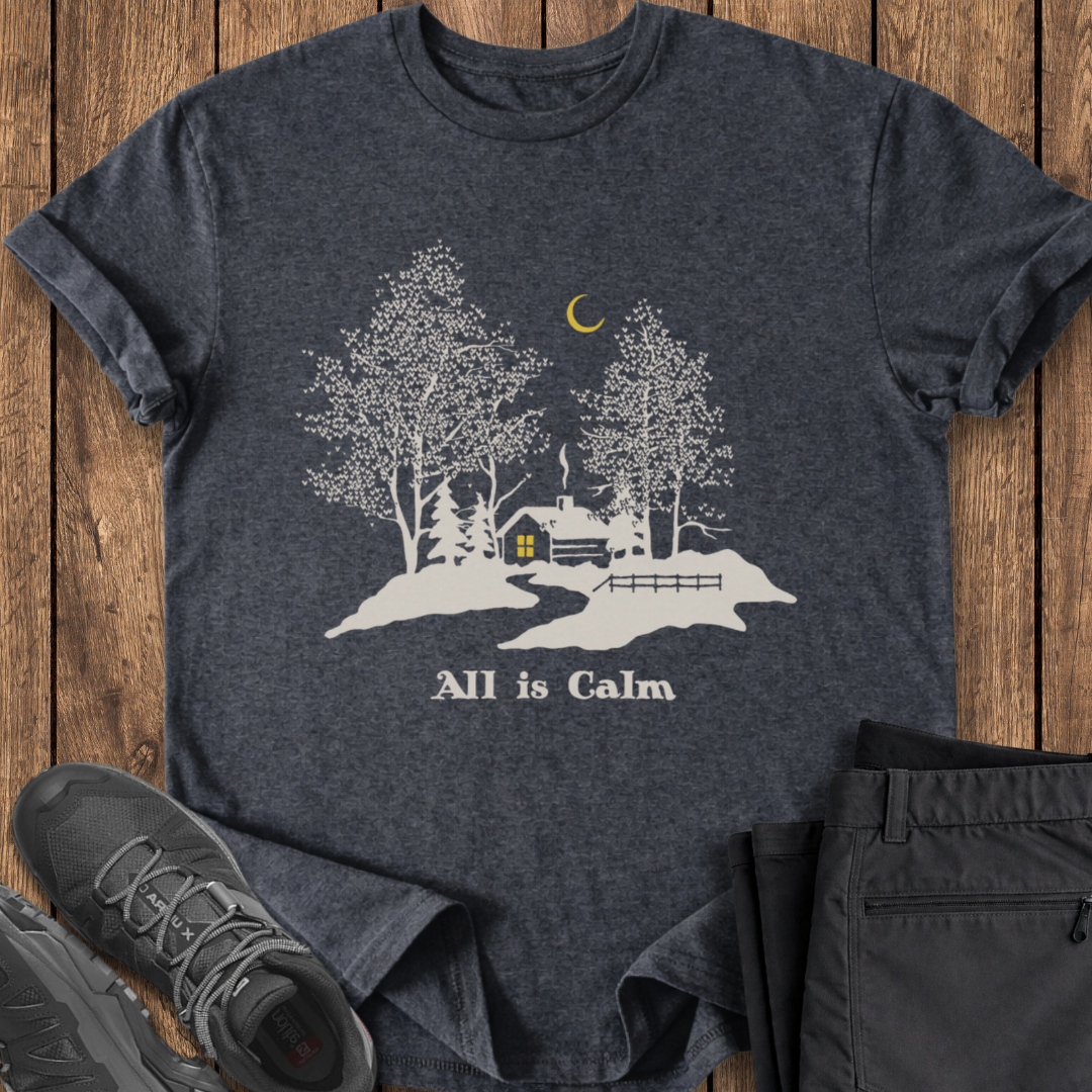 Dark gray t-shirt with a nature graphic and 'All is Calm' text on a wooden surface, with shoes and pants for scale. winter camp t shirt