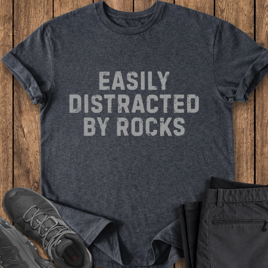 Dark gray t-shirt with 'EASILY DISTRACTED BY ROCKS' text on a wooden surface with shoes and pants.