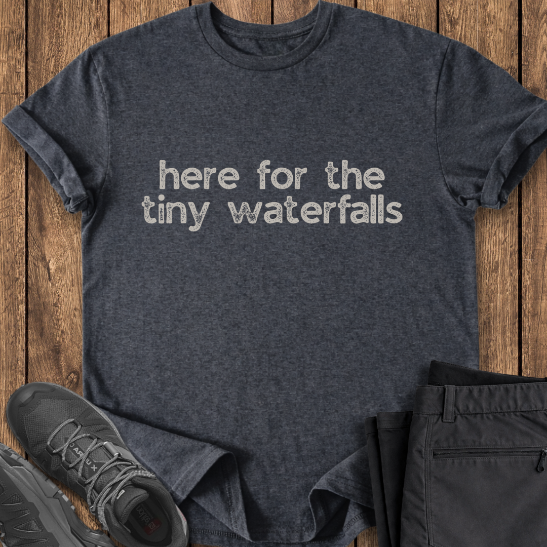 Dark gray t-shirt with 'here for the tiny waterfalls' text on a wooden surface, with shoes and pants partially visible. exploring hiking t shirt
