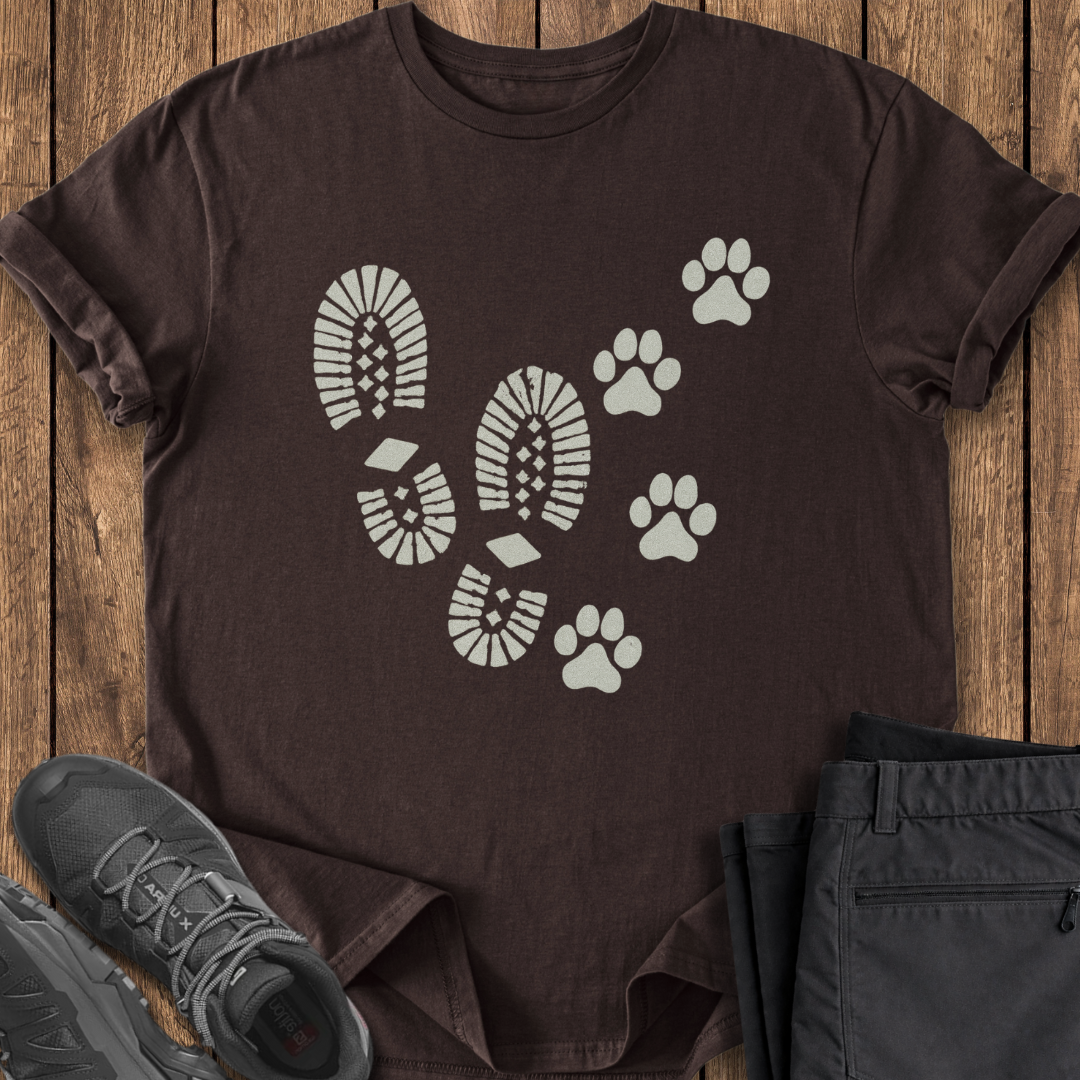 Brown t-shirt with white footprint and paw print design on a wooden surface, with black shoes and pants.