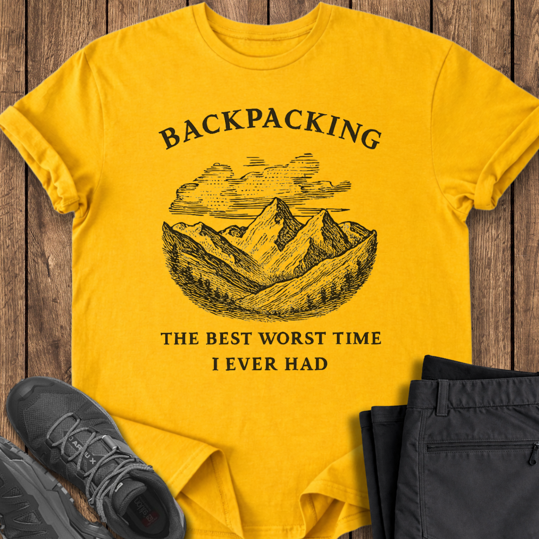 Yellow t-shirt with mountain graphic and text on a wooden surface with shoes and pants. funny backpacking t shirts