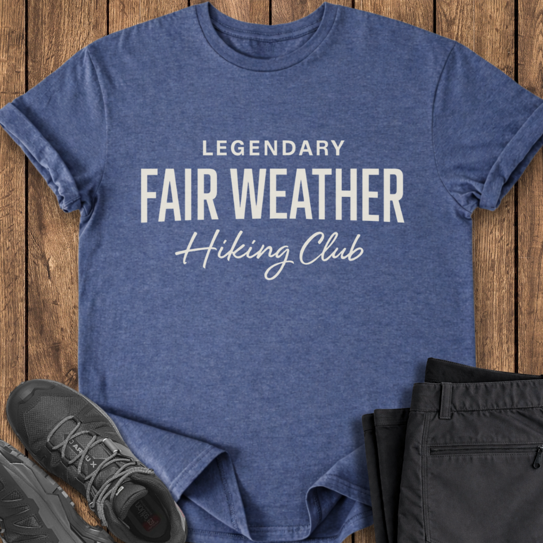Blue t-shirt with 'Legendary Fair Weather Hiking Club' text on a wooden surface with shoes and pants.