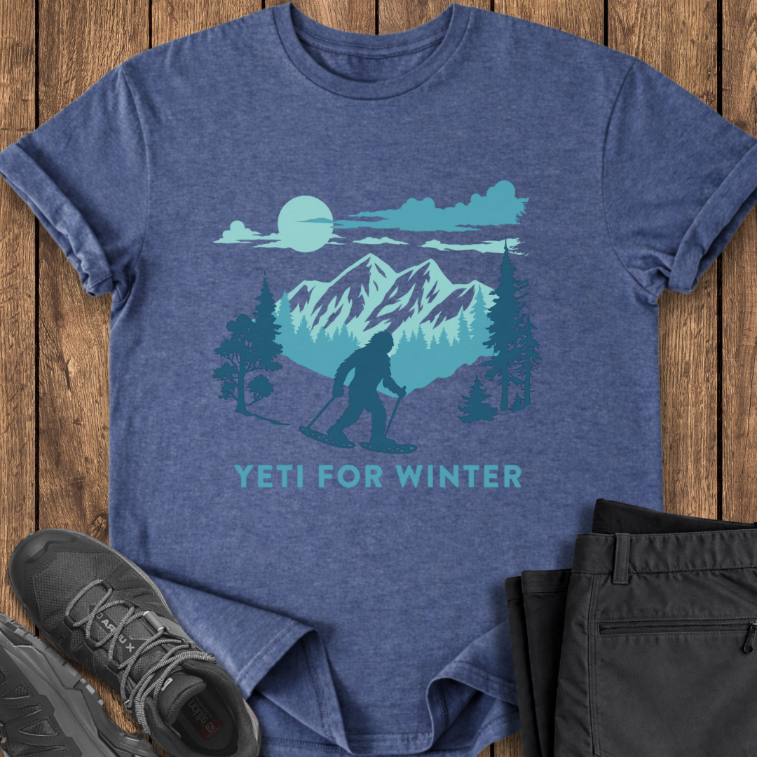 Blue t-shirt with mountain and sasquatch design, paired with black pants and shoes on a wooden surface.