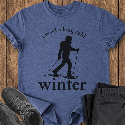 Blue t-shirt with skiing graphic and text on a wooden surface, with shoes and pants in the foreground. winter snowshoe snowshoeing t shirt